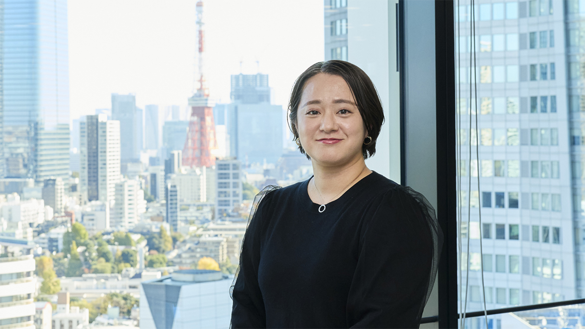 東京タワーが見えるオフィスの窓を背に、微笑む末永さんのポートレート写真。