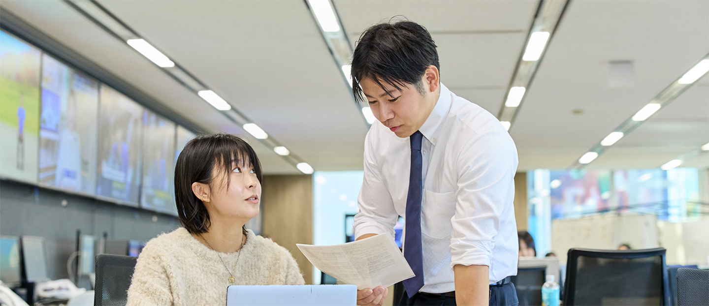 報道局のデスクで作業中の同僚に声をかけている八幡原さん。